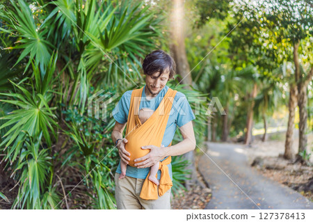 Father holding his little baby in a yellow sling in the park. Warm and loving family moment. Babywearing, fatherhood, and parent-child bonding concept 127378413