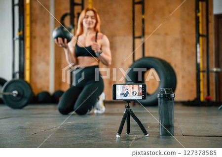 Smartphone is on the tripod, live stream, capturing video. Beautiful strong woman is in the gym 127378585