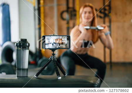 Doing squats, shooting video by smartphone. Beautiful strong woman is in the gym 127378602
