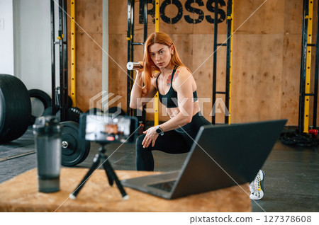 Laptop, bottle and smartphone on tripod is on the table. Beautiful strong woman is in the gym 127378608