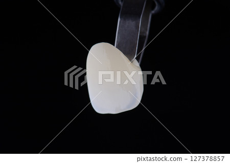 Close up shot ultra thin dental zirconium crown is held with tweezers with black background. 127378857