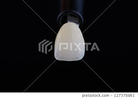 Close up shot ultra thin dental zirconium crown is held with tweezers with black background. 127378861