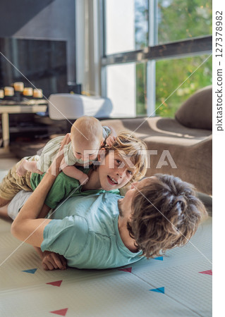 Father, son, and newborn baby lying on the floor in a cozy nursery, celebrating the arrival of their newest family member. Parenthood, bonding, and family love concept 127378982