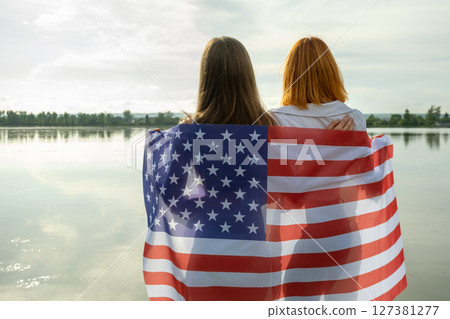 Two young friends women with USA national flag on their shoulders standing together outdoors on lake shore. Patriotic girls celebrating United States independence day. 127381277