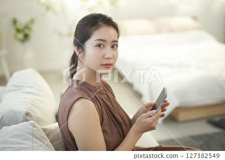 一位二十多歲的女性坐在沙發上操作智慧型手機 一位二十多歲的女性坐在沙發上操作智慧型手機 127382549
