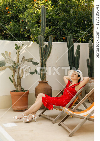 Relaxed woman in red dress sunbathing in lounge chair by cactus garden wall on a summer day Relaxed woman in red dress sunbathing in lounge chair by cactus garden wall on a summer day 127385041