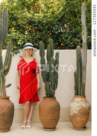 Woman in red dress and hat standing by cacti and orange trees on patio in warm summer daylight Woman in red dress and hat standing by cacti and orange trees on patio in warm summer daylight 127385042