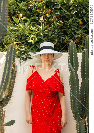 Stylish woman in red dress and wide hat standing near cactus and orange trees on sunny summer day Stylish woman in red dress and wide hat standing near cactus and orange trees on sunny summer day 127385045