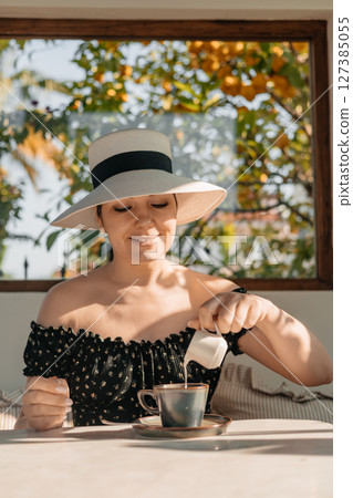 Woman in hat pouring milk into coffee at sunny morning breakfast table, enjoying peaceful moment Woman in hat pouring milk into coffee at sunny morning breakfast table, enjoying peaceful moment 127385055
