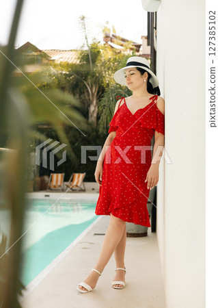 Elegant woman in red dress and hat standing by cactus, orange tree under sunlight in summer garden Elegant woman in red dress and hat standing by cactus, orange tree under sunlight in summer garden 127385102