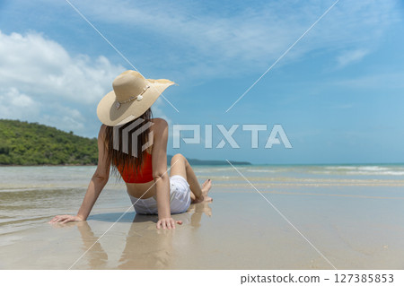 Women with straw hat, orange top and white shorts sitting on beach in tropical location with blue sky and turquoise sea. Back view. Women with straw hat, orange top and white shorts sitting on beach in tropical location with blue sky and turquoise sea. Back view. 127385853
