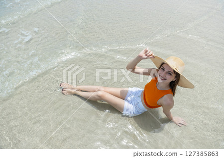 Young smiling women with straw hat, orange top and white shorts sitting on beach in tropical location with blue sky and turquoise sea. Young smiling women with straw hat, orange top and white shorts sitting on beach in tropical location with blue sky and turquoise sea. 127385863