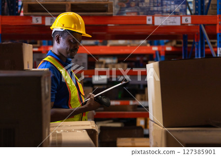 Man African American professional worker wearing safety uniform and white hard hat using bar code reader scanning box inspect product on shelves in warehouse. 127385901