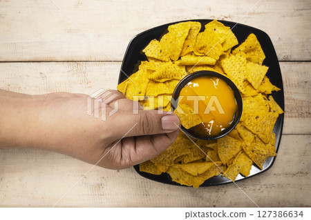 Human hand dipping a nachos tortilla chip in cheese sauce in a bowl on the table. Food and snacks concept 127386634