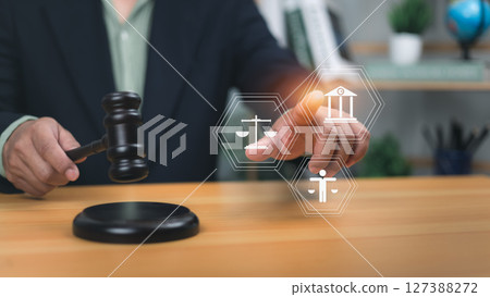 Human holding gavel pointing to holographic icons scales of justice, government building, and person. Symbolizes legal system, human rights, the rule of law for justice and regulatory compliance now. 127388272