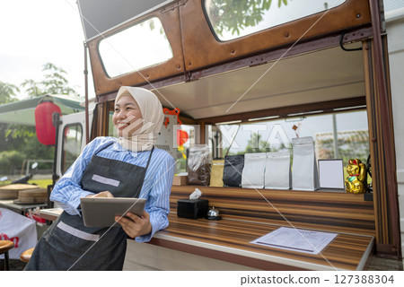 Indonesian southeast asian chinese muslim female barista in hijab standing while using a tablet in front of her coffee shop. A small business of a coffee shop or cafe on a truck 127388304