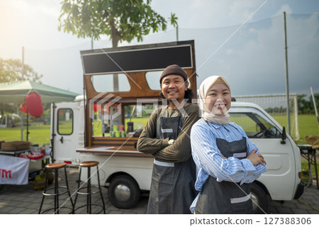 Indonesian southeast asian male and a Chinese muslim female in hijab couple barista standing with confidence in front of their coffee shop. A small business of a coffee shop or cafe on a truck 127388306