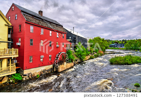 In Littleton, New Hampshire, United States, a red clapboard grist mill with a working waterwheel stands above the rushing Ammonoosuc River beneath a bright, cloud-dappled summer sky. 127388491