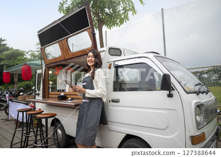 Indonesian southeast chinese asian female barista standing while holding a tablet in front of her coffee shop. A small business of a coffee shop or cafe on a truck 127388644