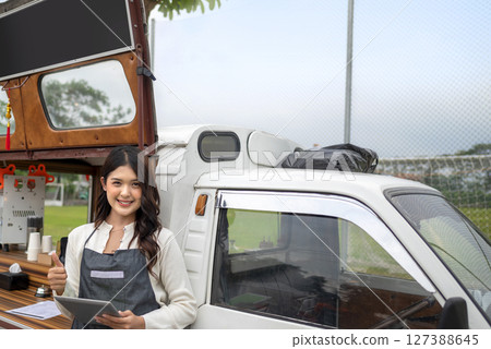 Indonesian southeast chinese asian female barista standing while holding a tablet and showing thumbs up gesture in front of her coffee shop. A small business of a coffee shop or cafe on a truck 127388645