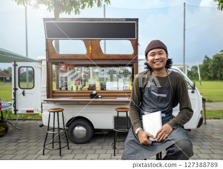 Indonesian southeast asian male barista sitting while holding a tablet in front of his coffee shop. A small business of a coffee shop or cafe on a truck 127388789
