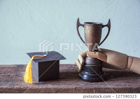 Graduation achievement trophy and cap on wooden background 127390323