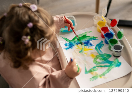 Young girl concentrated on her painting, sitting at a childrens table. Messy paint jars, vivid colors, and artistic expression. 127390340
