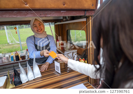 Indonesian southeast asian Chinese woman gives debit card to Chinese muslim female barista in hijab for payment after ordering. Mockup or copy space. Small business of a coffee shop or cafe on a truck Indonesian southeast asian Chinese woman gives debit card to Chinese muslim female barista in hijab for payment after ordering. Mockup or copy space. Small business of a coffee shop or cafe on a truck 127390761