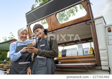 Indonesian southeast asian male and a Chinese muslim female in hijab couple of baristas using a cellphone in front of their coffee shop. A small business of a coffee shop or cafe on a truck 127391460