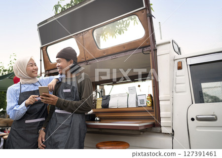 Indonesian southeast asian male and a Chinese muslim female in hijab couple of baristas using a cellphone in front of their coffee shop. A small business of a coffee shop or cafe on a truck 127391461