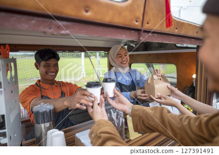 Indonesian southeast asian male and a Chinese muslim female in hijab couple baristas serving drinks and snacks to customers. A small business of a coffee shop or cafe on a truck Indonesian southeast asian male and a Chinese muslim female in hijab couple baristas serving drinks and snacks to customers. A small business of a coffee shop or cafe on a truck 127391491