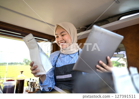 Indonesian southeast asian chinese muslim female barista in hijab sitting while using a tablet inside her coffee shop. A small business of a coffee shop or cafe on a truck 127391511