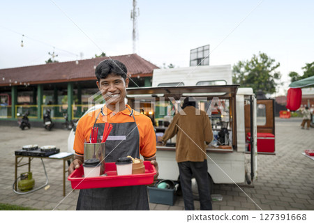 Indonesian southeast asian male barista holding a drink and food on a tray for serving to the customer. A small business of a coffee shop or cafe on a truck 127391668