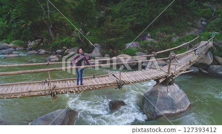 Aerial drone footage of a female tourist admiring the landscape from a precarious bamboo bridge built over a river flowing through a lush jungle 127393412