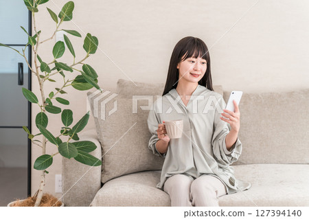 年輕的亞洲女性坐在家裡的沙發上喝咖啡，看著她的智慧型手機 127394140