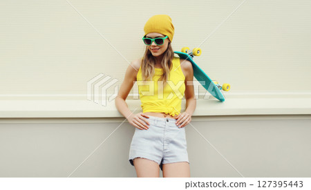 Stylish young woman with skateboard smiles, happy modern teenage girl posing on white wall 127395443