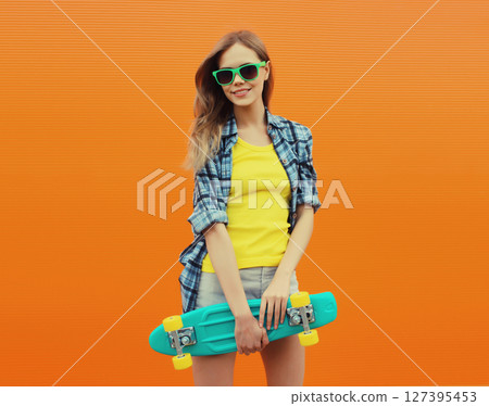 Stylish young woman with skateboard smiles, happy modern teenage girl posing on orange wall 127395453