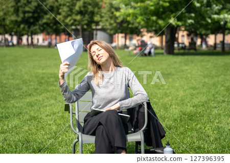 Pleased businesswoman fanning paperwork in hot weather sit in urban public space on lawn enjoy break 127396395