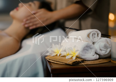 Spa scene with white rolled towels, frangipani flowers, and a blurred massage in the background 127396909