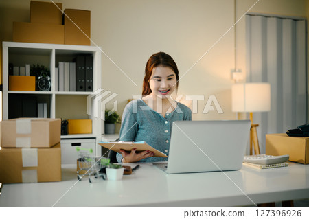 Female entrepreneur managing orders and working on laptop surrounded by shipping boxes 127396926