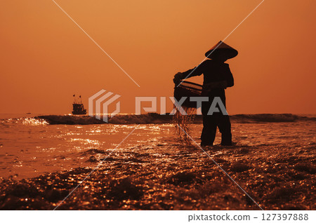 Silhouette of a Vietnamese woman in straw hat on shore of sea with ships in fishing village at sunset in Vietnam Silhouette of a Vietnamese woman in straw hat on shore of sea with ships in fishing village at sunset in Vietnam 127397888