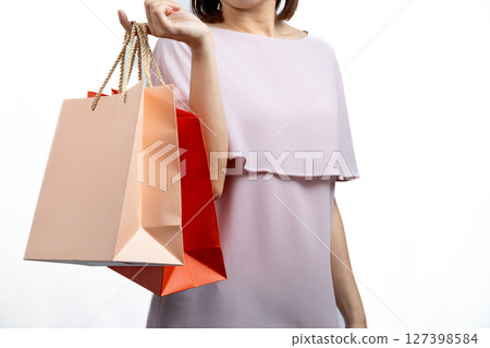 Woman in pastel pink dress holding orange and peach shopping bags with golden rope handles on her arm, isolated on white background; concept of retail or lifestyle spending 127398584