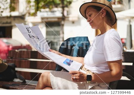 Smiling Woman Reading a Newspaper Outdoors in a Relaxed Urban Setting Smiling Woman Reading a Newspaper Outdoors in a Relaxed Urban Setting 127399446