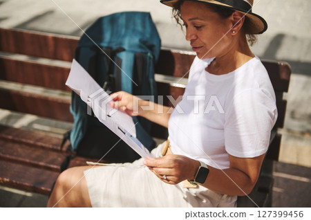 A woman sitting on a bench in a bright urban setting reads a map, exploring her surroundings. A woman sitting on a bench in a bright urban setting reads a map, exploring her surroundings. 127399456
