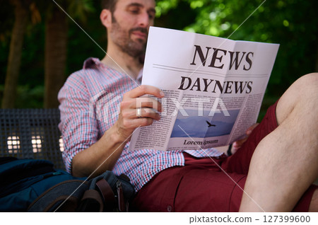 Man Sitting Outdoors Reading a Newspaper in the Park Surrounded by Green Trees 127399600