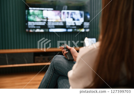 Woman is sitting at home watching TV, holding remote controller 127399703