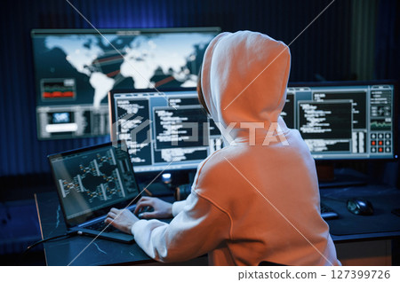 In white hoodie. Young professional female hacker is indoors by computer with lot of information on displays 127399726