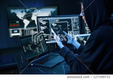 Black colored hoodie. Young professional female hacker is indoors by computer with lot of information on displays Black colored hoodie. Young professional female hacker is indoors by computer with lot of information on displays 127399731