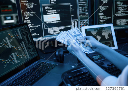 With currency in hands. Young professional female hacker is indoors by computer with lot of information on displays With currency in hands. Young professional female hacker is indoors by computer with lot of information on displays 127399761