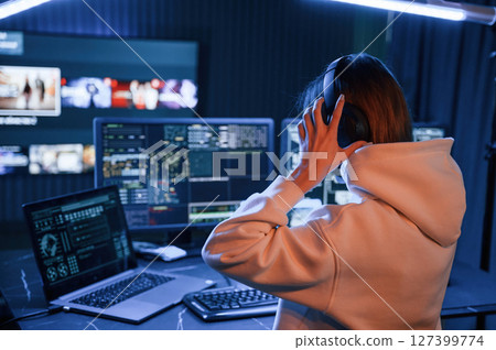 Wearing headphones. Young professional female hacker is indoors by computer with lot of information on displays 127399774